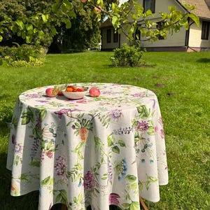 Op de afbeelding: Een rond tafelkleed met een botanische print van diverse kleurrijke bloemen, groene bladeren en stengels op een lichtbeige achtergrond. Een kleine witte kom met rode appels en klein fruit staat in het midden op het oppervlak van de stof.