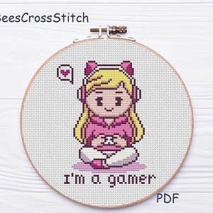 May include: Cross-stitch embroidery in a wooden hoop featuring a pixelated image of a person with blonde hair wearing a pink hoodie and headphones, holding a game controller. The text "I'm a gamer" is below the image, with a heart icon. The brand name "BeesCrossStitch" is at the top.