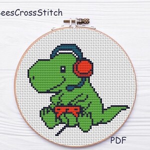 May include: Cross-stitch embroidery of a green dinosaur wearing a headset and holding a red controller. The dinosaur is centered in a wooden embroidery hoop against a white background. The text "BeesCrossStitch" is at the top, and "PDF" is at the bottom.
