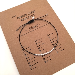 Könnte beinhalten: Ein schwarzes Armband aus Schnur mit silbernen Perlen, die das Wort "Schwester" in Morsecode buchstabieren. Das Armband ist auf einer Karte mit einer Morsecode-Alphabet-Tabelle abgebildet.