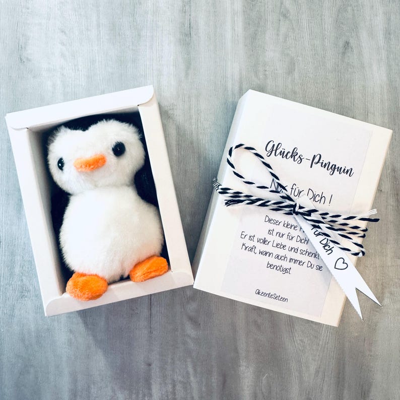 May include: A white gift box with a small white penguin plush toy inside. The box has a white tag with black text that reads "Gl&uuml;cks-Pinguin No fur Dich!" and a smaller white tag with black text that reads "Tur Dich!" with a black heart.