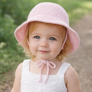 Könnte beinhalten: Ein hellrosa gehäkelter Sonnenhut mit breiter Krempe und Kinnbändern. Der Hut wird von einem kleinen Kind mit blonden Haaren und blauen Augen getragen. Das Kind trägt ein weißes Kleid.