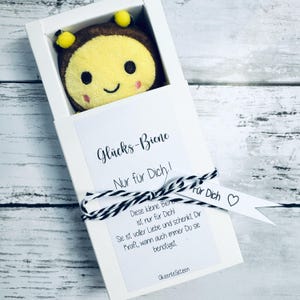 May include: A plush bee toy with a smiling face is presented in a white box. The box has a tag with the text "Gl&uuml;cks-Biene Nur f&uuml;r Dich!" and a black and white string tied around it. The bee is yellow, brown, and pink.