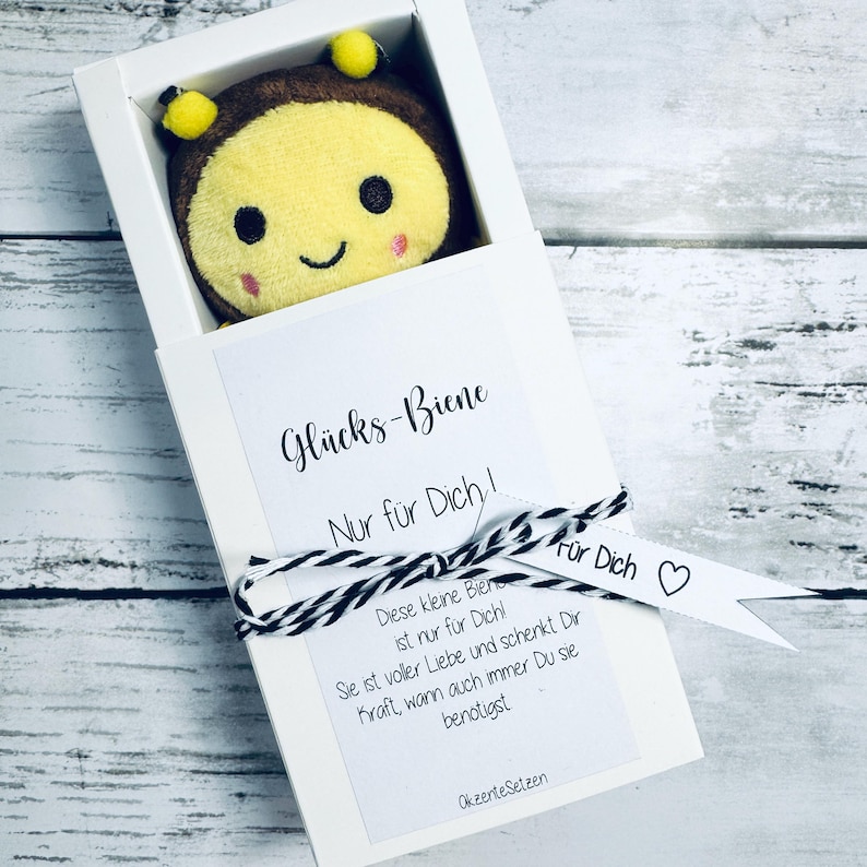 May include: A plush bee toy with a smiling face is presented in a white box. The box has a tag with the text "Gl&uuml;cks-Biene Nur f&uuml;r Dich!" and a black and white string tied around it. The bee is yellow, brown, and pink.