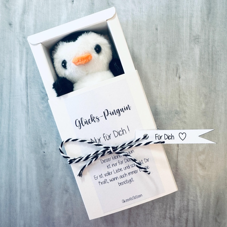 May include: A white gift box with a black and white penguin plush toy inside. The box has a white tag with the text "Gl&uuml;cks-Pinguin Nur f&uuml;r Dich!" and a white tag with the text "F&uuml;r Dich" with a heart symbol.