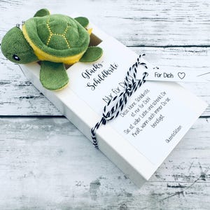 Könnte beinhalten: Ein grünes und gelbes Plüschtier in Form einer Schildkröte liegt auf einer weißen Geschenkbox, die mit schwarz-weißem Garn gebunden ist. Auf der Box steht deutscher Text, darunter "Glücks-Schildkröte" und "Nur für Dich". Ein kleines Etikett trägt die Aufschrift "Für Dich".