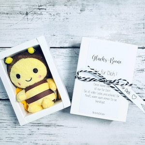 May include: A plush bee toy in a white box, next to a white card tied with black and white twine. The bee is yellow, brown, and black. The card has text in German, including "Gl&uuml;cks-Biene" and "F&uuml;r Dich!"