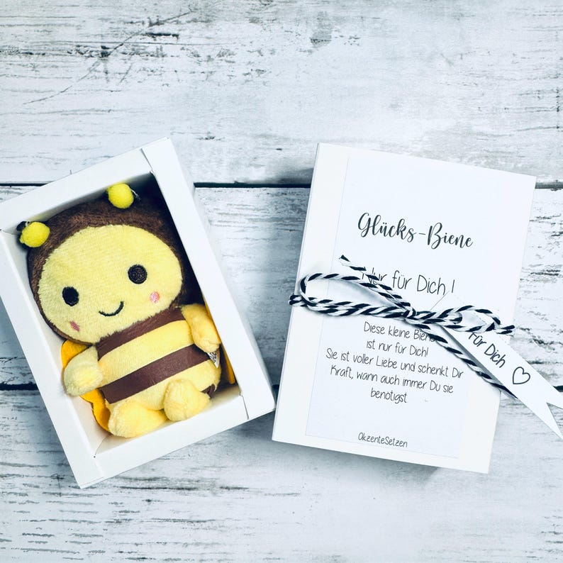 May include: A plush bee toy in a white box, next to a white card tied with black and white twine. The bee is yellow, brown, and black. The card has text in German, including "Gl&uuml;cks-Biene" and "F&uuml;r Dich!"
