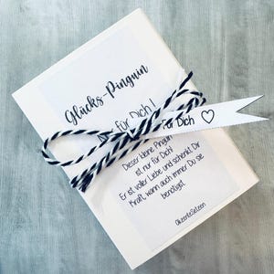 May include: A white gift box with a black and white striped ribbon tied around it. The box has a white tag attached to it with the text "Gl&uuml;cks-Pinguin" and "f&uuml;r Dich!" in black. The tag also has a small heart symbol and the text "Dieser kleine Pinguin ist nur f&uuml;r Dich! Er ist voller Liebe und schenkt Dir Kraft, wann auch immer Du sie ben&ouml;tigst." in black.