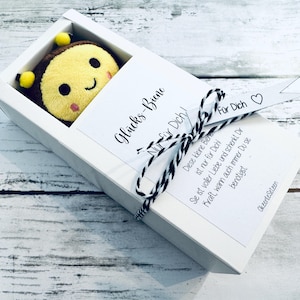 May include: A plush bee toy with a smiling face, presented in a white gift box. The box is tied with a black and white string and a ribbon that says "For You". The box also has text in German.