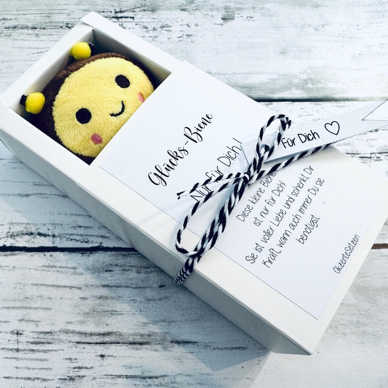 May include: A plush bee toy with a smiling face, presented in a white gift box. The box is tied with a black and white string and a ribbon that says "For You". The box also has text in German.