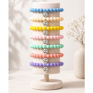Könnte beinhalten: Eine Präsentation von Armbändern mit bunten Perlen in Blau, Creme, Gelb, Rosa, Mintgrün, Koralle und Lila. Jedes Armband hat einen kleinen Silberanhänger mit der Aufschrift "LOVE & PEACE". Die Armbänder sind auf einem weißen Ständer angeordnet.