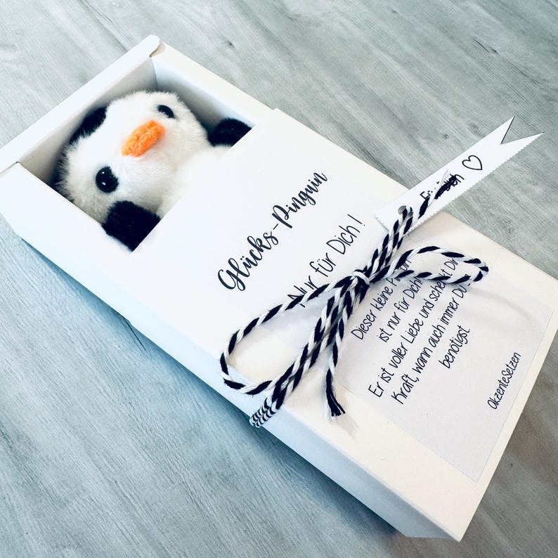 May include: A white box with a black and white ribbon tied around it. The box contains a small stuffed penguin and a card that reads "Gl&uuml;cks-Pinguin nur f&uuml;r Dich!"  The card also has a message that says "Dieser kleine Freund ist nur f&uuml;r Dich. Er ist voller Liebe und schenkt Dir Kraft, wann auch immer Du ben&ouml;tigst."