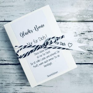 May include: A white card with the text "Gl&uuml;cks-Biene" and "Nur f&uuml;r Dich!" in black script. The card is tied with black and white twine and has a small white tag that says "F&uuml;r Dich" with a heart. The card also has additional text in German.