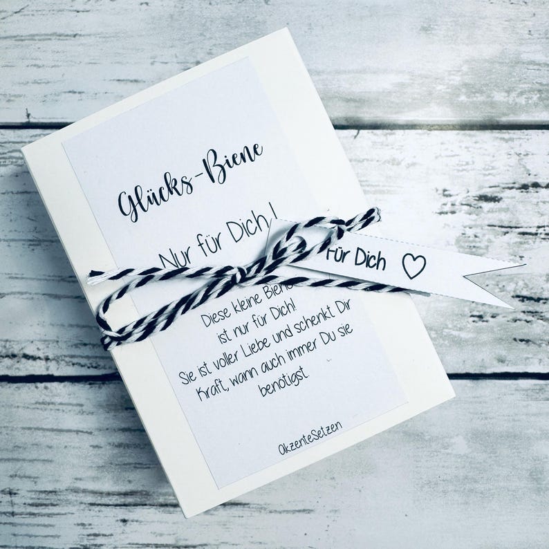 May include: A white card with the text "Gl&uuml;cks-Biene" and "Nur f&uuml;r Dich!" in black script. The card is tied with black and white twine and has a small white tag that says "F&uuml;r Dich" with a heart. The card also has additional text in German.