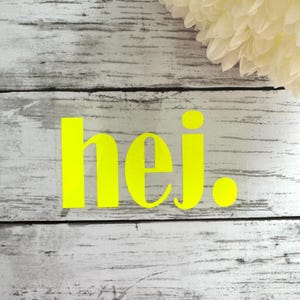 Könnte beinhalten: Ein neon-gelber Vinyl-Aufkleber mit dem Wort "hej" auf einem weißen Holzgrund.