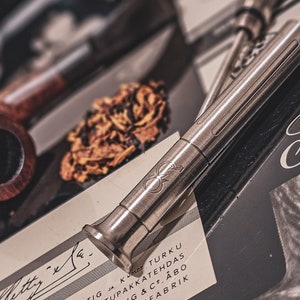 Könnte beinhalten: Eine silberne Metallpfeife mit glatter, polierter Oberfläche. Die Pfeife hat ein abgerundetes Ende und einen langen, schlanken Körper. Die Pfeife liegt auf einem Stück Papier mit Schrift darauf. Die Schrift ist in einer kursiven Schriftart und wird teilweise von der Pfeife verdeckt.