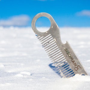 Könnte beinhalten: Ein silberner Metallkamm mit einem Flaschenöffner und einem Loch im Griff. Der Kamm liegt im Schnee. Der Kamm hat den Text "SISUMAN&" auf der Seite.