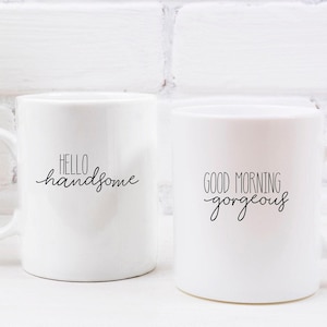 Op de afbeelding: Twee witte keramische mokken met zwarte tekst. De ene mok zegt "Hello Handsome" en de andere mok zegt "Good Morning Gorgeous".