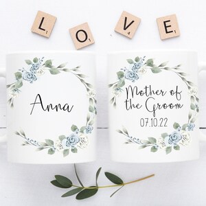Può includere: Due tazze in ceramica bianca con disegni floreali blu e verdi. Una tazza dice "Anna" e l'altra dice "Madre dello sposo 07.10.22".