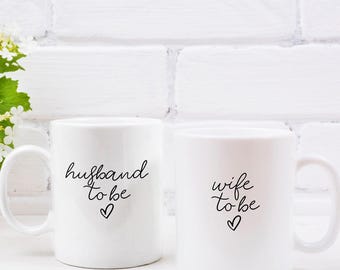 Può includere: Due tazze in ceramica bianca con testo nero. La tazza a sinistra dice "husband to be" con un cuore. La tazza a destra dice "wife to be" con un cuore.