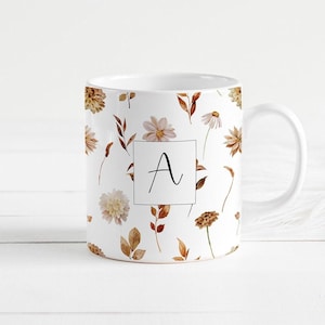 Peut inclure: Une tasse en céramique blanche avec un motif floral dans des tons de brun et beige. La tasse a une grande lettre "A" au centre.