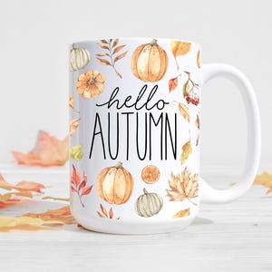 Könnte beinhalten: Weiße Keramiktasse mit Herbstmotiv. Die Tasse zeigt ein Muster aus Kürbissen, Blättern und Beeren. Der Text "hello autumn" ist in Schwarz auf die Tasse gedruckt.