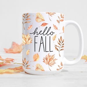 Könnte beinhalten: Weiße Keramiktasse mit einem schwarz-weißen "hello fall" Text und einem Muster aus braunen und orangefarbenen Aquarellblättern.