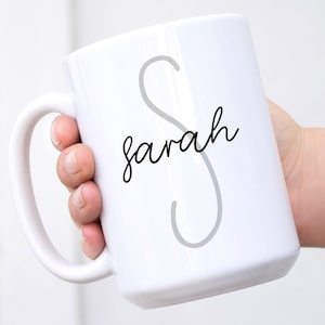 Pode incluir: Caneca de cerâmica branca com uma letra "S" cursiva cinza e o nome "sarah" escrito em cursiva preta.