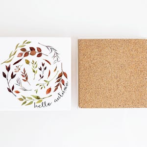 May include: Ceramic coaster with a white background and a circle of watercolour leaves in shades of green, brown, and red. The text "hello autumn" is written in black cursive inside the circle. The coaster has a cork backing.
