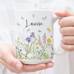 May include: White ceramic mug with a floral design featuring butterflies, bunnies, and flowers. The mug has the name "Louise" written on it.