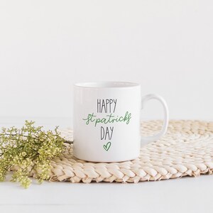 May include: White ceramic mug with the text "HAPPY St. Patrick's DAY" in black and green script, with a green heart below. The mug sits on a woven placemat with a sprig of greenery.