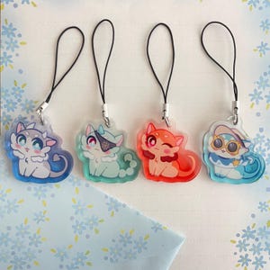 May include: Four colorful cat-shaped acrylic charms with black lanyards. Each charm features a unique cat design with different colors and accessories, including an eye patch and sunglasses. The charms are on a light blue floral background.