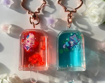 May include: Two clear plastic keychain charms shaped like popsicles with a gold metal ring. The charms contain a red and blue liquid with a small cartoon character inside each.