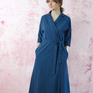 Può includere: Un lungo abito a portafoglio di colore blu scuro. L'abito presenta un colletto, maniche a tre quarti e una cintura in tessuto abbinata che si stringe in vita. Le tasche sono visibili sulla parte anteriore dell'abito. Lo sfondo è un muro strutturato rosa e bianco.