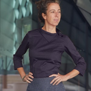 Könnte beinhalten: Eine Frau trägt eine dunkelviolette, langärmelige Bluse mit Kragen und einem Knopfdetail an der Schulter. Sie trägt außerdem einen dunkelblauen Rock mit einem weißen geometrischen Muster.