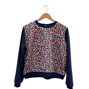 Può includere: Una felpa blu navy con un motivo leopardato arancione e bianco sul davanti. Le maniche e lo scollo sono blu navy tinta unita. La felpa è appesa a una gruccia di legno.