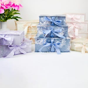 May include: An assortment of decorative boxes in different sizes and colours. The boxes feature floral patterns and are tied with satin ribbons in shades of blue, lavender, and pink. Some boxes are stacked, others are arranged separately.
