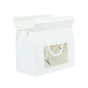 May include: White cardboard box with a clear window and a white rope handle. The box is designed for packaging and displaying baked goods.