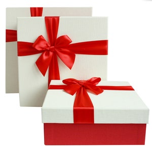 May include: Three square gift boxes with white textured lids and red bases. Each box is tied with a vibrant red satin ribbon and bow. The boxes are arranged in a layered composition, showcasing the gift-wrapping.