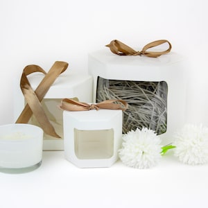 Puede incluir: Tres cajas de regalo hexagonales blancas con ventanas transparentes, la caja más grande está llena de papel triturado y atada con una cinta marrón, la caja mediana está vacía y la caja más pequeña contiene una vela blanca, todas las cajas tienen lazos de cinta marrón.