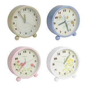 May include: Four round analog alarm clocks in pastel colors: beige, blue, pink, and white. Each clock features a floral design on the face and small ball feet. The clock hands are black, and the numbers are clearly visible.
