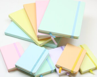 May include: A collection of pastel-colored notebooks with matching elastic bands and ribbon bookmarks. The notebooks are in shades of pink, blue, green, yellow, and orange. The notebooks are stacked and arranged on a white surface.