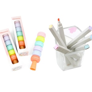 Peut inclure: Ensemble de fournitures artistiques colorées, comprenant des marqueurs en forme de macarons et un ensemble de marqueurs aux couleurs pastel dans un récipient transparent. L'emballage indique "Macaron". Les marqueurs sont dans des tons orange, jaune, vert, bleu et violet.