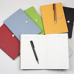May include: An open notebook with lined pages and a black pen, surrounded by several closed notebooks in various colors: red, blue, green, yellow, and black. Each notebook has a matching elastic closure.