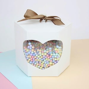 May include: White hexagonal gift box with a heart-shaped window filled with pastel-coloured spheres. The box is tied with a brown ribbon. The box is on a pastel background.