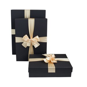 Puede incluir: Tres cajas de regalo negras apiladas una encima de la otra. Cada caja está atada con una cinta de satén de color marfil ancha y un lazo.
