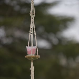 Puede incluir: Un macetero de macramé con un anillo de madera y una vela rosa dentro de un frasco de vidrio. El macetero está suspendido de una rama de árbol.
