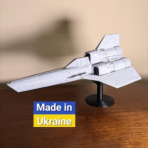 May include: A white 3D printed model of a spaceship on a black stand. The model has a long, narrow body with a pointed nose and two large engines at the rear. The text "Made in Ukraine" is printed on a yellow and blue background below the model.