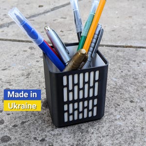 May include: A black plastic pen holder with a geometric design. The holder is filled with various pens, including a blue pen, a silver pen, a green pen, a yellow pen, and a black pen. The holder is sitting on a gray concrete surface. Made in Ukraine.